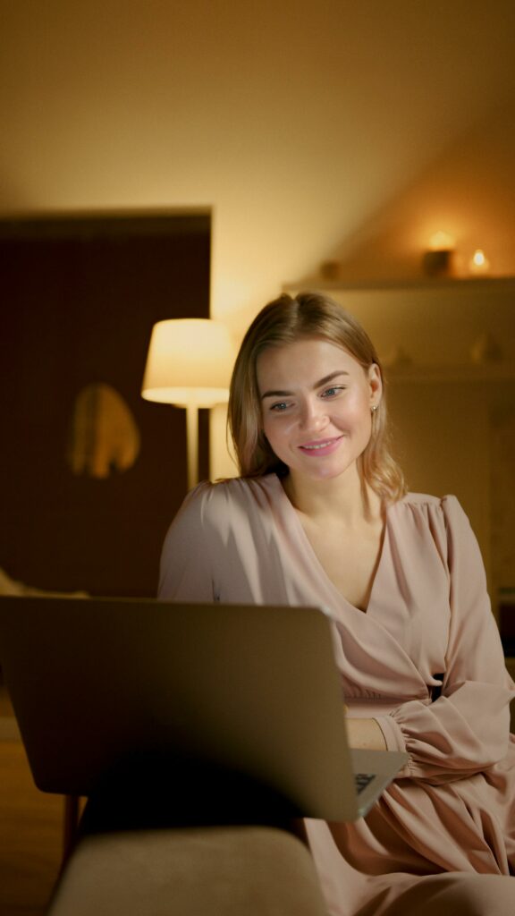 A young woman enjoys browsing on her laptop in a comfortable home setting with warm lighting.